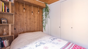 Un dormitorio con cabecera de madera, cama con colcha estampada, estantería con libros y una planta colgante.