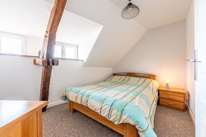 Ein Schlafzimmer mit schrägem Dach, einem Bett mit gestreifter Bettdecke, einem Nachttisch aus Holz mit einer Lampe und einem großen Fenster.