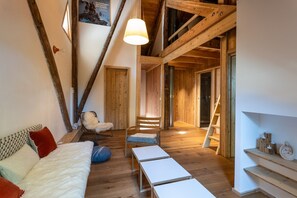 Avoin, loft-tyylinen huone, jossa on näkyvillä puiset kattopalkit, valkoinen sohva, tuoli ja tikapuut, jotka johtavat ylemmälle tasolle.
