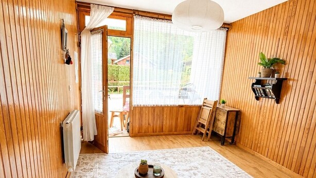 Et hyggeligt rum med trævægge, et rundt bord med en plante, en sofa med mønstrede puder og en reol.