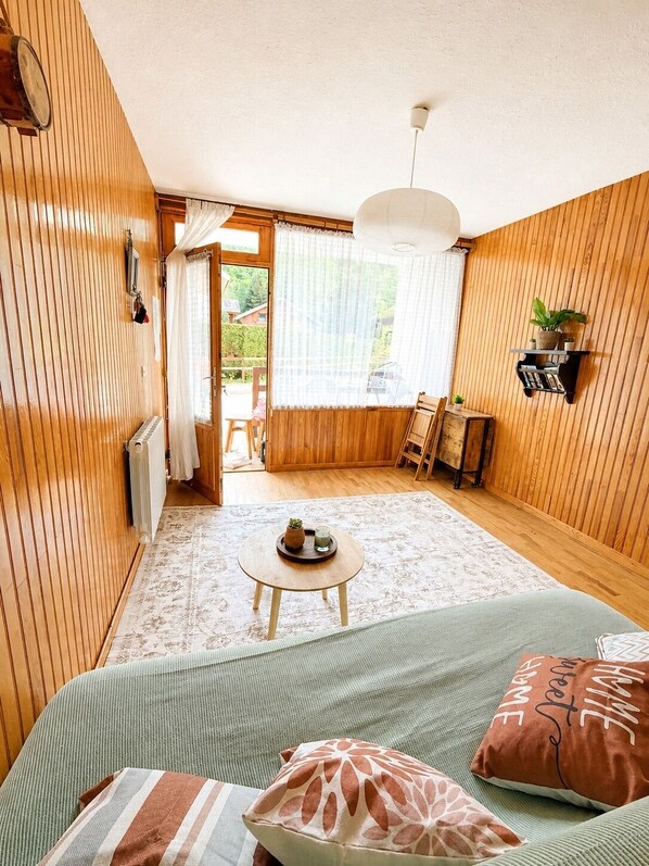 Ein gemütliches Zimmer mit Holzwänden, einem runden Tisch mit Pflanze, einer Couch mit gemusterten Kissen und einem Bücherregal.