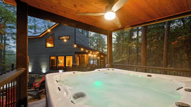 A hot tub with a wooden deck, a house with string lights, and a forested backdrop. - Pine Hill Lodge - Hot Tub, Fire Pit, Sleeps 14 (Broken Bow)