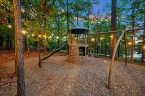 A playground with a slide and climbing structures, surrounded by trees and a gravel surface. - Pine Hill Lodge - Hot Tub, Fire Pit, Sleeps 14 (Broken Bow)