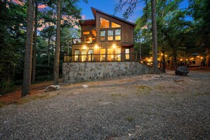 A two-story wooden cabin with stone walls, surrounded by trees and a gravel driveway. - Pine Hill Lodge - Hot Tub, Fire Pit, Sleeps 14 (Broken Bow)