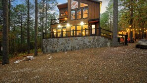 A modern wooden cabin with a stone foundation, large windows, and a balcony overlooking a forested area. - Pine Hill Lodge - Hot Tub, Fire Pit, Sleeps 14 (Broken Bow)