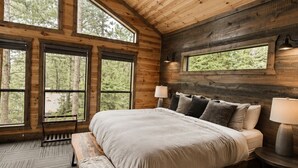 A wooden cabin bedroom with a large bed, a ceiling fan, and multiple windows offering a view of the forest. - Pine Hill Lodge - Hot Tub, Fire Pit, Sleeps 14 (Broken Bow)
