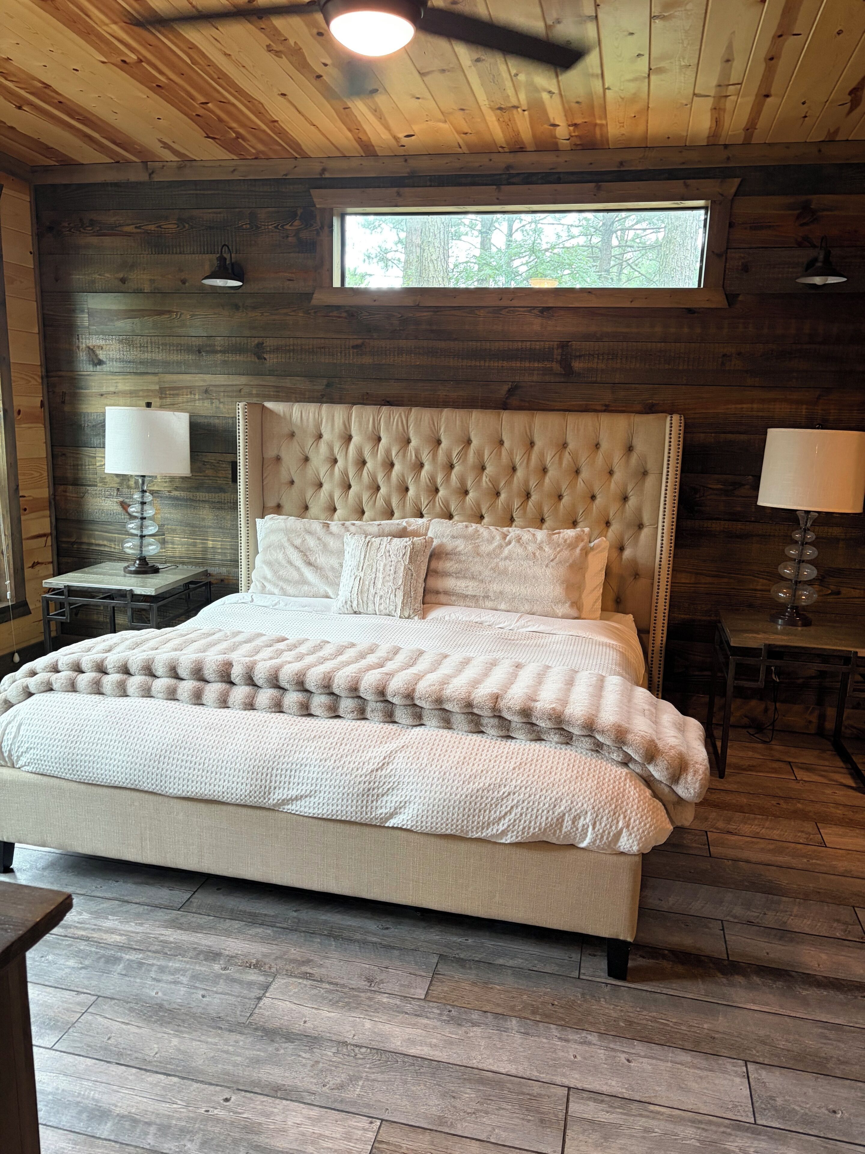 Une chambre à coucher avec une tête de lit capitonnée, une grande fenêtre, un plancher en bois et deux lampes de chevet.