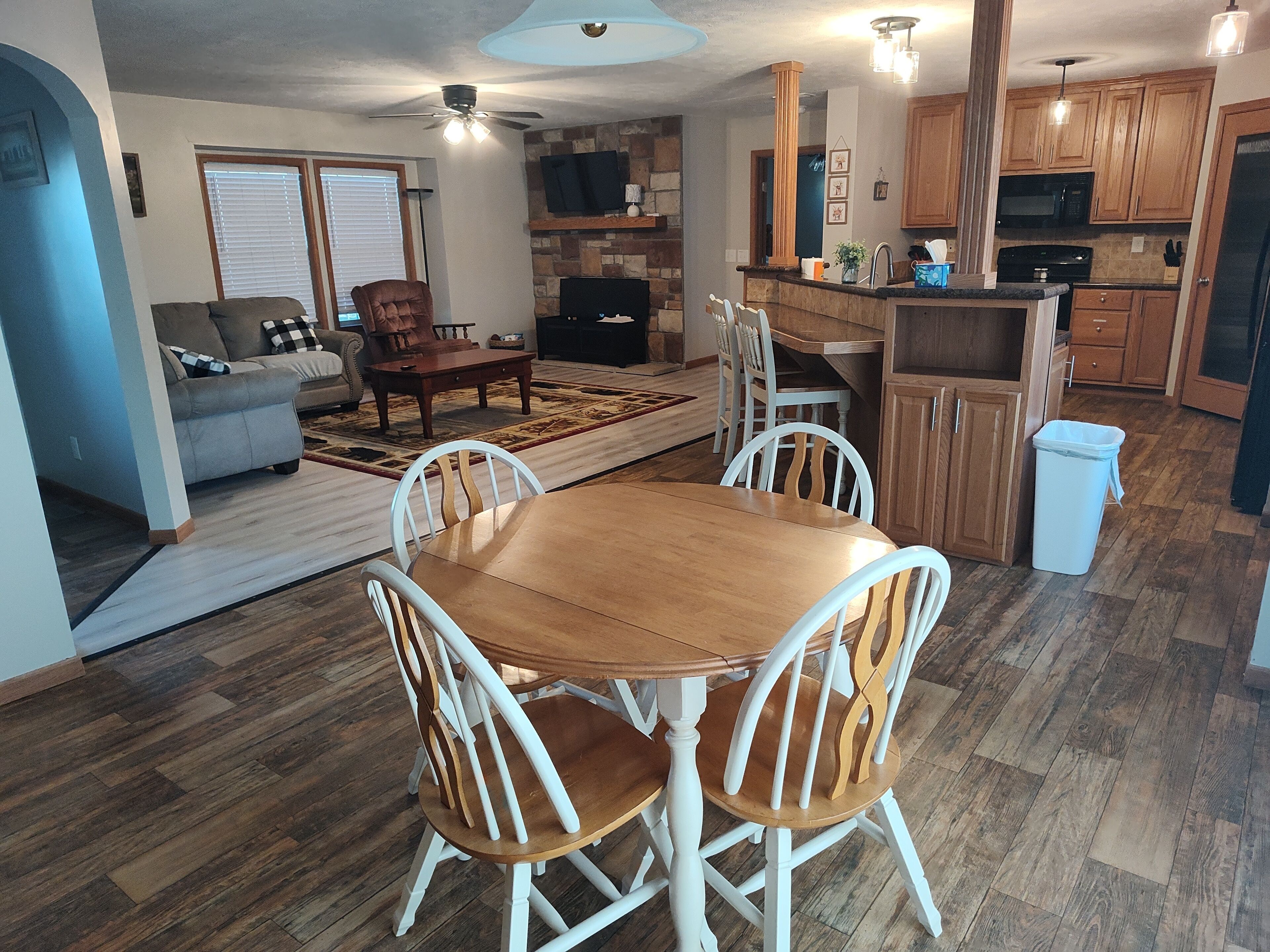 A dining area with a wooden table and white chairs, a kitchen with wooden cabinets, a living room with a sofa and a fireplace, and a ceiling fan.