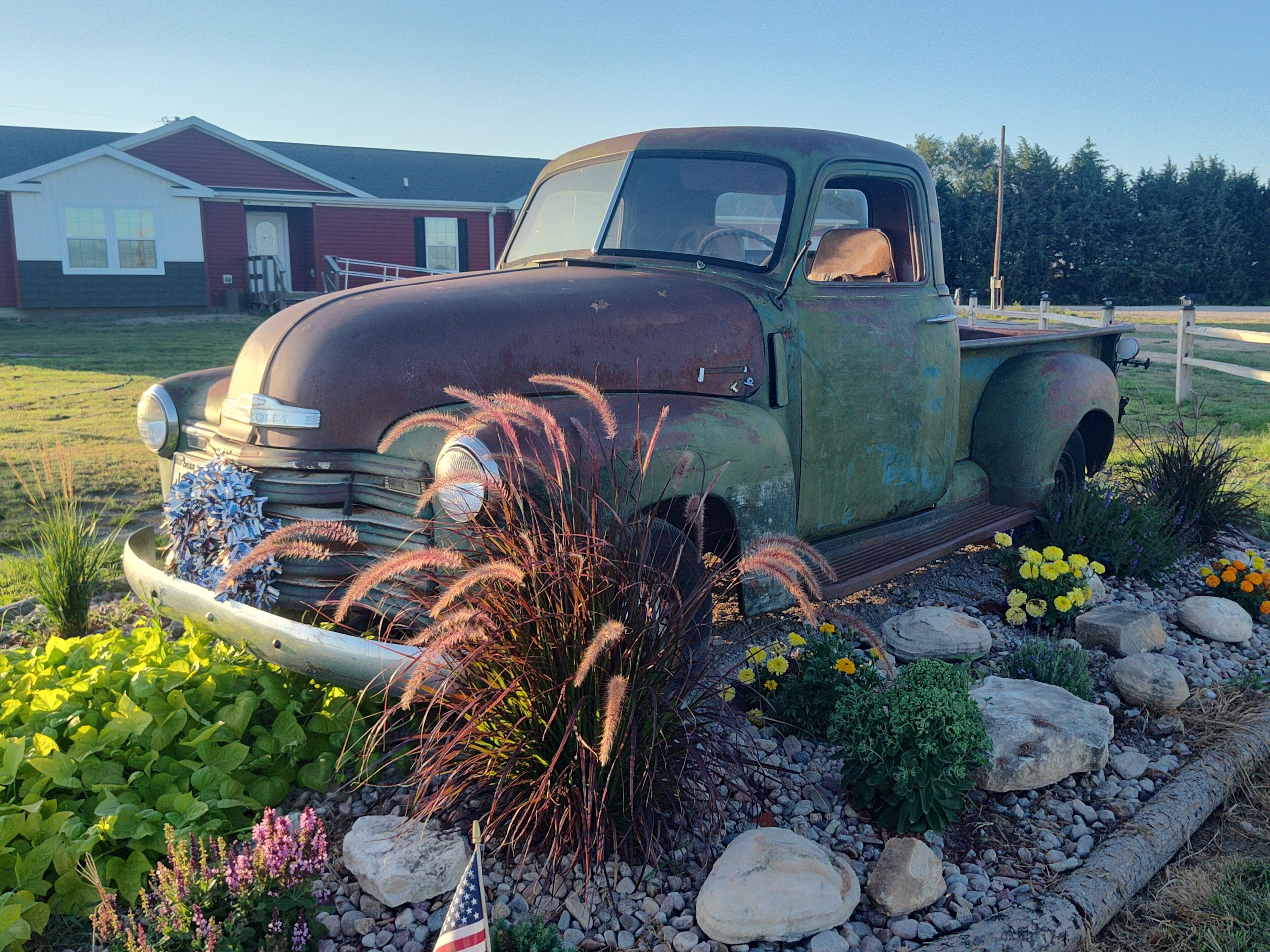 An old truck parked in a garden with rocks, plants, and a flag.