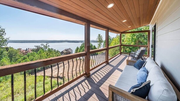 A covered outdoor deck with a wooden railing, a cushioned seating area, and a view of a lake and surrounding greenery. - Enjoy stunning Pickwick Lake views and water access from Aqua Vista Pickwick! (Hardin County)