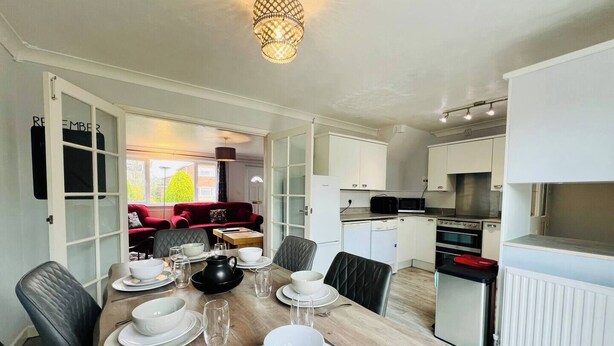 A modern kitchen with white cabinets, a dining table set for four, and a red sofa in the background. - Sociable Suffolk Home | A12, Ipswich & Colchester (Capel Saint Mary)