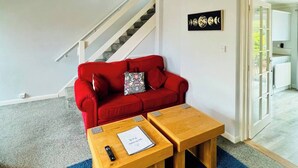 A modern living room with a red sofa, two wooden side tables, a remote control, and a welcome pack on the sofa. - Sociable Suffolk Home | A12, Ipswich & Colchester (Capel Saint Mary)