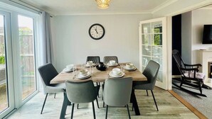 A dining area with a wooden table, four chairs, a wall clock, and a television on the wall. - Sociable Suffolk Home | A12, Ipswich & Colchester (Capel Saint Mary)
