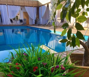 Una piscina con bordo in legno, tende bianche e sedie a sdraio.