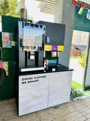 A coffee vending machine with a 'DRINK COFFEE BE SMART' sign, located outdoors with a green wall and a building in the background. - Hotel7 (Kapchagay)