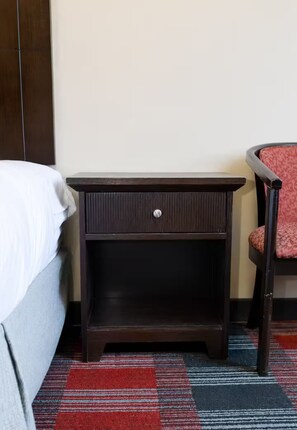 Een donker houten nachtkastje met een lade, een rode fauteuil met een patroon en een bed met wit beddengoed.