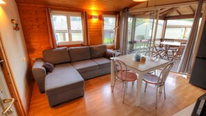 A cozy room with a wooden ceiling, a gray sofa, a dining table with chairs, and a view through large windows. - Vacation Rental in Cap d'Agde: "LES PESCALUNES" (Cap d'Agde)