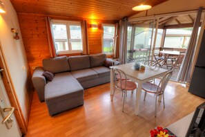A cozy room with a wooden ceiling, a gray sofa, a dining table with chairs, and a view through large windows.