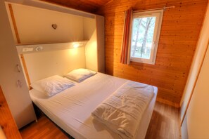 A single bed with white linens in a cabin-style room with wooden walls and a window with curtains.