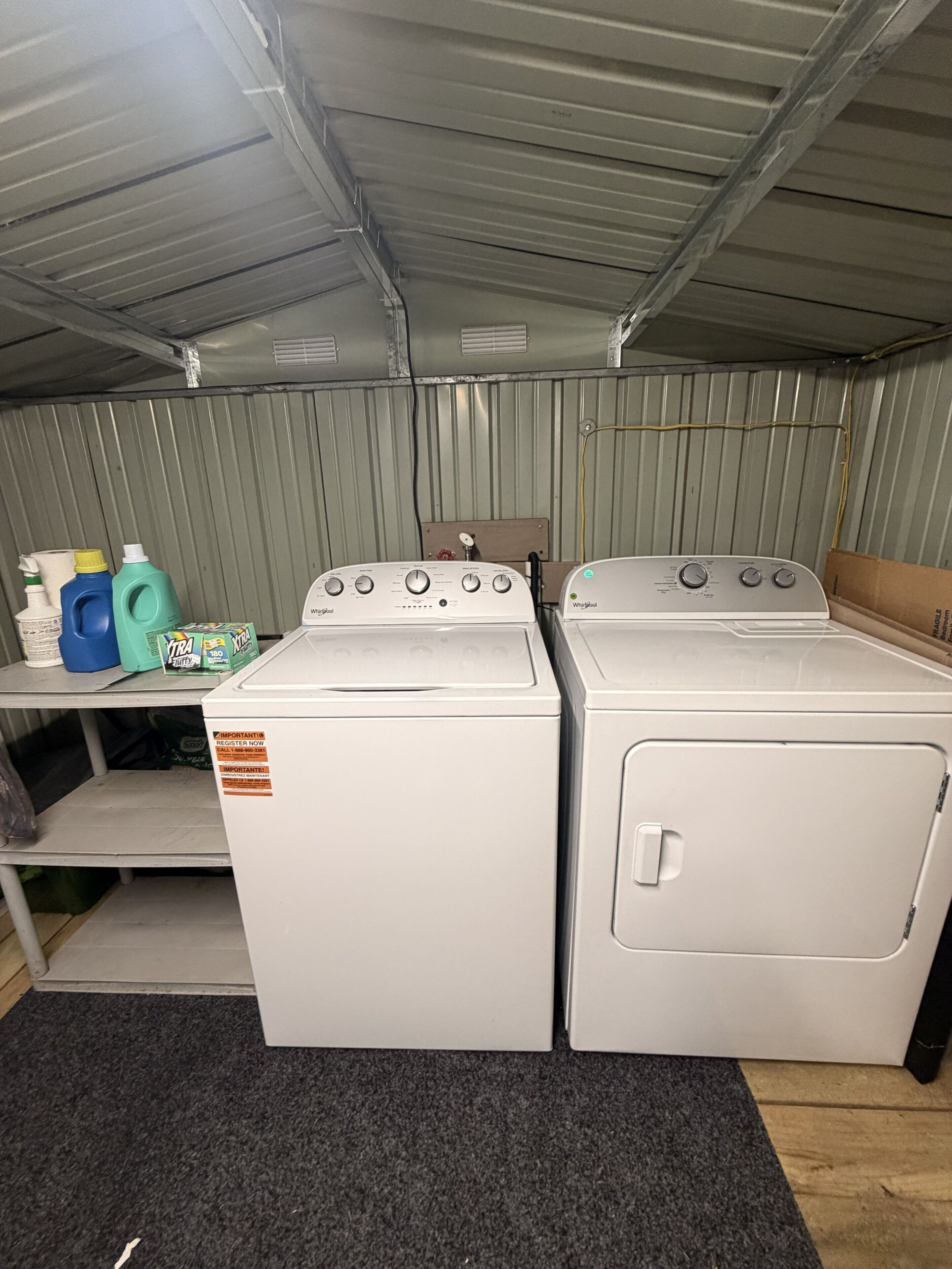 Une buanderie équipée de deux machines à laver et deux sèche-linge Whirlpool blancs, d’une étagère métallique et de produits de nettoyage.