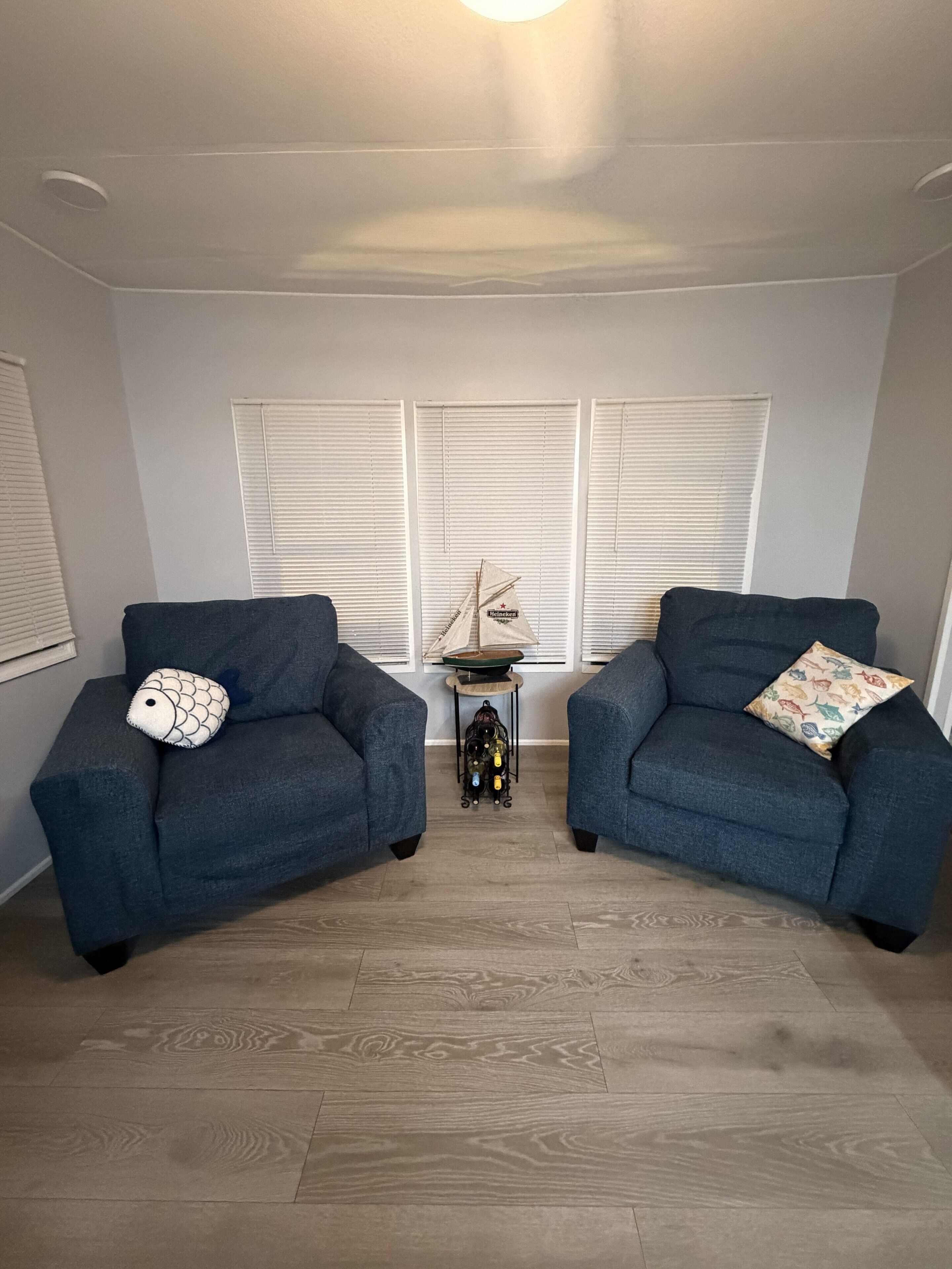 Una habitació amb dues butaques blau fosc, una taula auxiliar amb una ampolla i un objecte decoratiu, i una finestra amb persianes.