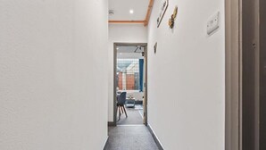 En smal hall med ett heltäckningsmatta på golvet, en dörr som leder till ett rum med en stol och ett bord, och en vägg med en ljusströmbrytare och en liten prydnadsföremål.