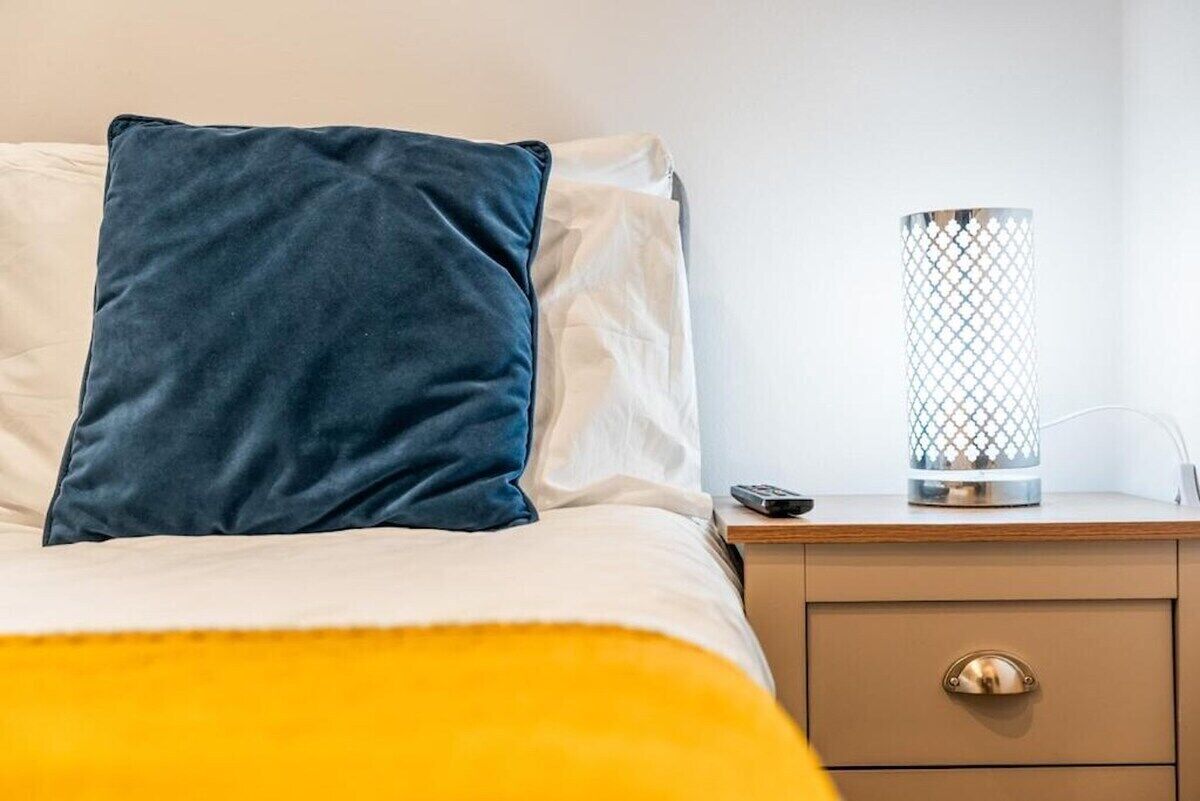 A bed with a dark-colored pillow, a white pillow, and a yellow blanket. There is a bedside table with a lamp and a remote control.