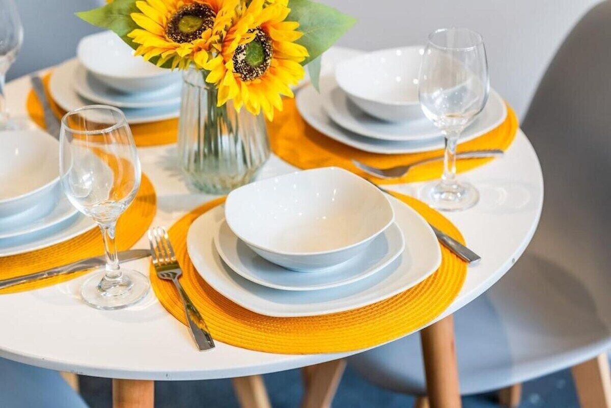 Une table à manger dressée avec de l’assiette blanche, des bols et des couverts, agrémentée d’un vase rempli de tournesols.