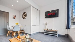 Ett modernt vardagsrum med en matplats, en platt-TV som visar ”NETFLIX”, en klocka och en bänk.