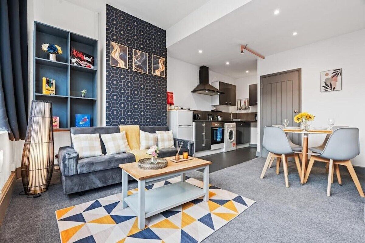 A modern living room with a sofa, coffee table, and dining area with chairs. There is a kitchen in the background with a washing machine and a dining table with a vase of flowers.