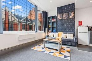 A modern living room with a sofa, coffee table, and a patterned rug. There is a bookshelf and a kitchen area visible. - Stylish City-Centre Stay in Leicester | Market St (Leicester)