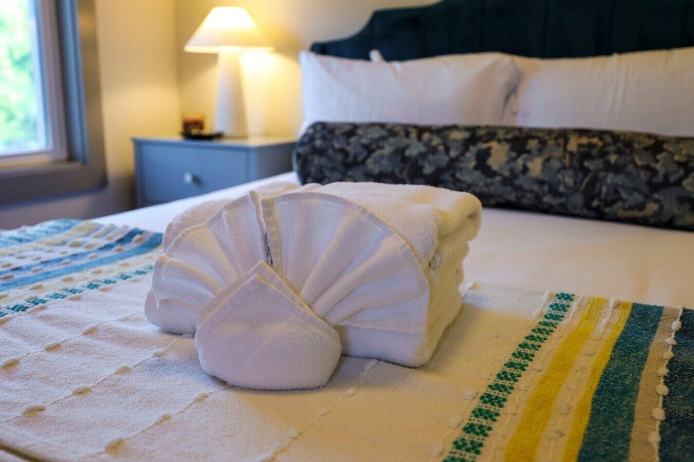 Een opgemaakt bed met een gevouwen handdoek, een lamp op een nachtkastje en een raam met daglicht.
