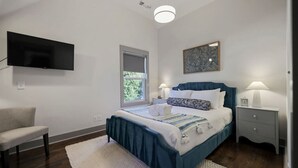 Ein Schlafzimmer mit einem Bett, einem Sessel, einem Nachttisch, einer Lampe, einer Uhr und einem an der Wand befestigten Fernseher.
