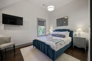 Ein Schlafzimmer mit einem Bett, einem Sessel, einem Nachttisch, einer Lampe, einer Uhr und einem an der Wand befestigten Fernseher.