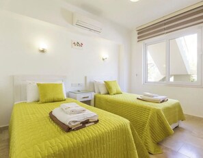 Ein Hotelzimmer mit zwei Betten, jeweils mit einer grünen Bettdecke und Kissen, einem Nachttisch mit einer Lampe und einem Fenster mit Jalousien.