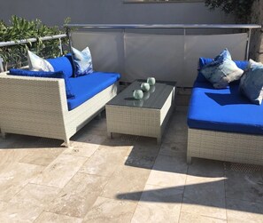 Een terras met twee rieten banken en een bijzettafeltje, voorzien van een glazen blad en decoratieve vazen.
