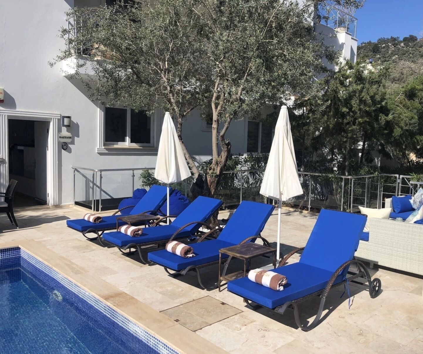 Un espace aménagé au bord de la piscine, avec des chaises longues et des parasols, une piscine à l’eau d’un bleu limpide et un bâtiment à la façade blanche.