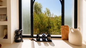 A kitchen with a coffee maker, three teapots, and a pitcher on the counter, with a window overlooking greenery. - Palais Pleya (Essaouira)