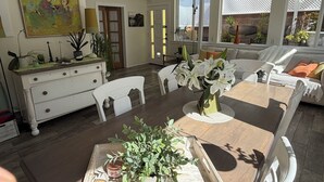 En rummelig opholdsstue med spisebord, hvide stole, sofa og flere planter.
