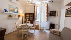 A dining area with a round table, chairs, a cabinet, and a window with curtains. - Albis 32 - Two Bedroom Apartment, Sleeps 4 (Alghero)