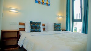 A hotel room with a bed, a painting, two bedside lamps, a window with curtains, and a wooden nightstand. - 1 Bedroom Blue Lagoon Resort Suite with Private Balcony in Paje (Paje)