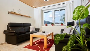 Ein modernes Wohnzimmer mit einem schwarzen Sofa, einem Holztisch und einem großen Fenster mit Blick ins Freie.