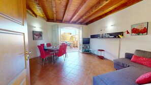 A living room with a sofa, a dining area with chairs, a TV stand, and a kitchen area with a counter. - Borgo Torchio F23 by Bookinggardalake (Manerba del Garda)