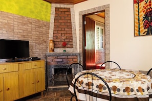 Una habitación con chimenea de ladrillo, un televisor sobre un mueble de madera, una mesa de comedor con mantel estampado y un cuadro en la pared.