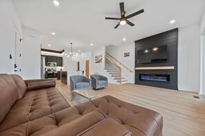 Ein modernes Wohnzimmer mit einer braunen Ledersofa, zwei grauen Sesseln, einem Glastisch, einem Kamin, einem Fernseher und einer Treppe, die zu einer oberen Ebene führt.