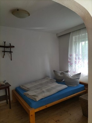 Une petite chambre avec un lit simple, un cadre en bois, un matelas bleu et une fenêtre ornée de rideaux à motifs floraux.