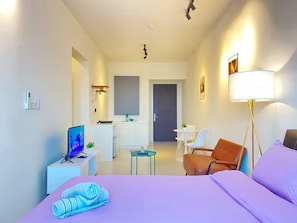 A modern hotel room with a bed, a desk, a chair, a small table, and a kitchenette.