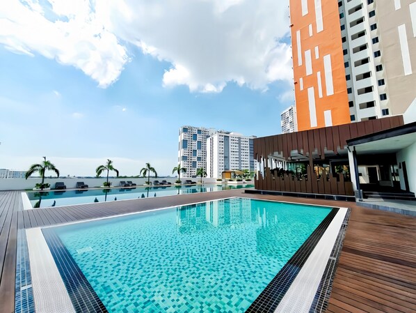 A rooftop swimming pool with a wooden deck, surrounded by high-rise buildings and palm trees.
