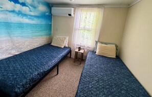 A small room with two beds, a window with curtains, a wall-mounted air conditioner, and a small table with a lamp.