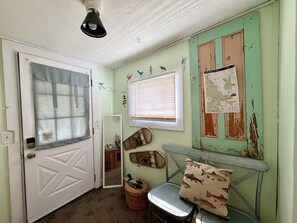 A room with a white door, a window with blinds, a mirror, a chair, a basket, and a wall with a map and bird decorations.